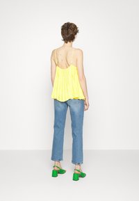 Top amarelo de pregas sem mangas, de corte solto, com alças finas. Combinado com jeans de perna reta azul-claro e sandálias verdes de salto bloco.