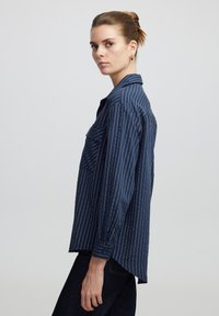 Marineblauwe lange mouwen shirt met witte verticale strepen, knoopsluiting aan de voorkant, een enkele borstzak en een licht gebogen zoom.