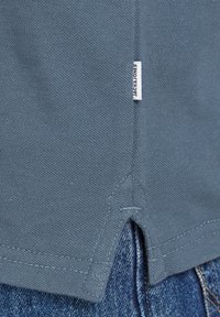 Modrá bavlněná tričko má štítek se značkou "JACK & JONES." Měkký zaoblený lem je kombinován s džínovou tkaninou níže.