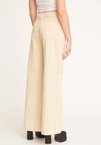 Pantaloni beige a gamba larga con vita alta, dotati di due tasche posteriori, frontale piatto e dettagli di cucitura. Indossati con scarpe con tacco plateau.