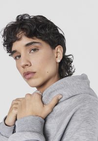 Felpa grigia con cappuccio e polsini a costine, indossata da una persona. Orecchini e anelli a cerchio argentati completano l'outfit, con trucco naturale e capelli texturizzati.