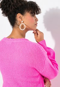 Maglione rosa a maglia con un motivo testurizzato e a costine, e un ampio scollo. La modella indossa grandi orecchini a cerchio con dettagli in perle.