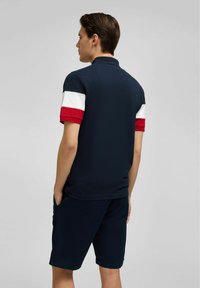 Marineblaues Poloshirt mit rot-weiß-roten gestreiften Ärmeln; kombiniert mit dunklen Shorts; kurze Ärmel und lockerer Schnitt.
