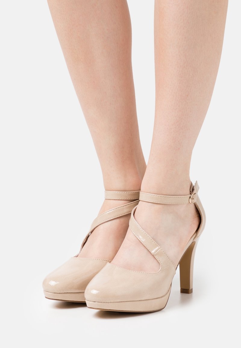 Chaussures à talons hauts en cuir verni beige avec brides à la cheville, portées pieds nus sur un fond blanc uni.