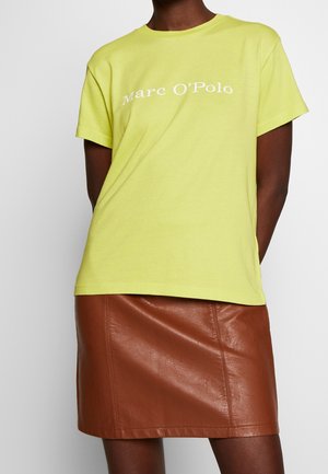 Osoba ubrana w jasnozieloną koszulkę z krótkim rękawem z napisem "Marc O’Polo" oraz brązową skórzaną spódnicę na jednolitym tle.