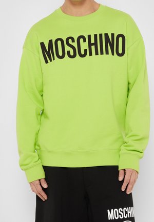 Person iført limegrøn sweatshirt med sort "MOSCHINO" tekst over brystet og sorte bukser med hvid "MOSCHINO" tekst.