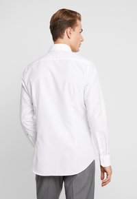 Witte langemouwen shirt gezien van de achterkant, met een klassieke kraag, op maat gesneden pasvorm en enkele knopen bij de manchetten. Gladde textuur.