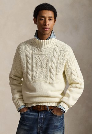 Άνδρας φοράει πουλόβερ με πλεκτά καλώδια σε κρεμ χρώμα με υφή λογοτύπου, τζιν πουκάμισο από κάτω, κρυμμένο μέσα σε μπλε τζιν με καφέ ζώνη, χέρια στις τσέπες.