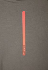 Šedé tričko z látky s vertikálním červeným pruhem a názvem značky „Kappa“ v bílé barvě na spodní části. Měkká textura, neformální design.