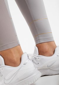 Jasnoszare legginsy z złotymi poziomymi paskami, z teksturowanym elementem. Połączone z białymi butami sportowymi.
