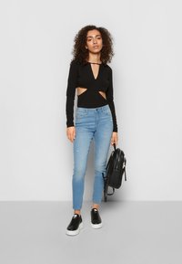 Body de manga longa preto com recortes laterais, combinado com jeans justos azul claro. Usa ténis pretos e carrega uma mochila preta.