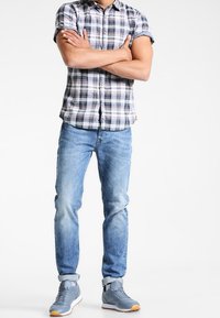 Camisa de cuadros de manga corta en tonos azul, blanco y verde, combinada con vaqueros ajustados azul claro y zapatillas grises con suelas blancas y detalles naranjas.