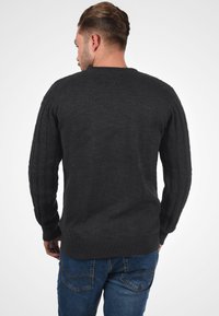 Suéter de punto gris oscuro con un cuello acanalado y detalles de punto de cable a lo largo de las mangas, combinado con jeans azules.