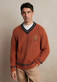 Pomarańczowy sweter o teksturowanej powierzchni z dekoltem w serek i granatowymi paskami. Posiada emblemat z literą "B" na klatce piersiowej oraz detale na mankietach. Nosi się go na białej koszuli.