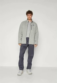 Pilkos vilnos striukės su priekiniais užtrauktukais ir šoninėmis kišenėmis, derinamos su tamsiai mėlynomis kelnėmis ir šviesia avalyne, prieš neutralų foną.