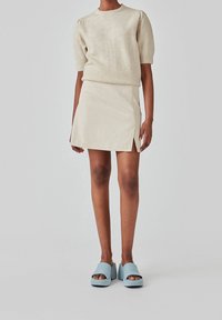 Pull en tricot beige à manches courtes, coupe ajustée. Mini jupe beige clair avec fentes latérales. Sandales à talons épais bleu avec bout ouvert.