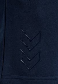 Donkerblauwe stof met een gestructureerd, ribbelachtig oppervlak. Heeft een matte chevronlogo dat nabij de onderkant is ingebed, wat het ontwerp versterkt.