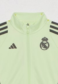 Chaqueta con cremallera en verde claro con franjas negras, con logotipo y marca del equipo bordados. Tejido suave y diseño atlético.