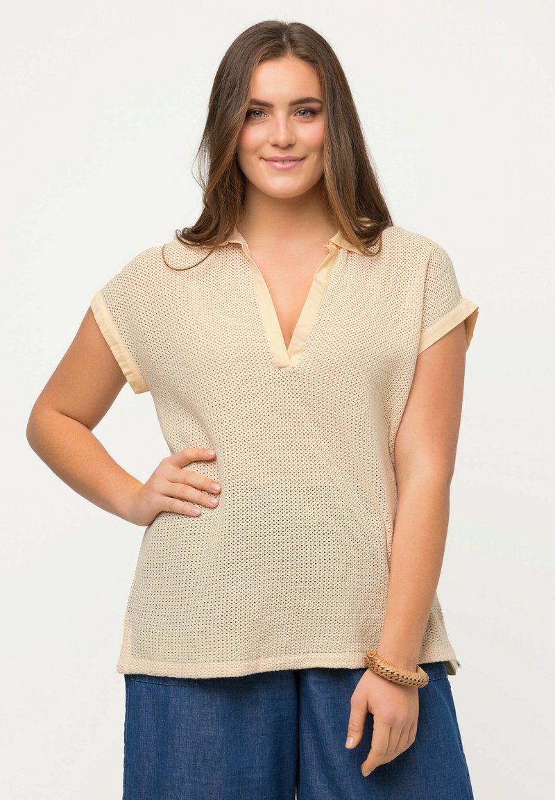 Top corto a maniche corte in maglia beige chiaro con scollo a V e orli arrotolati, caratterizzato da un motivo testurizzato e fessure laterali per facilitare i movimenti.