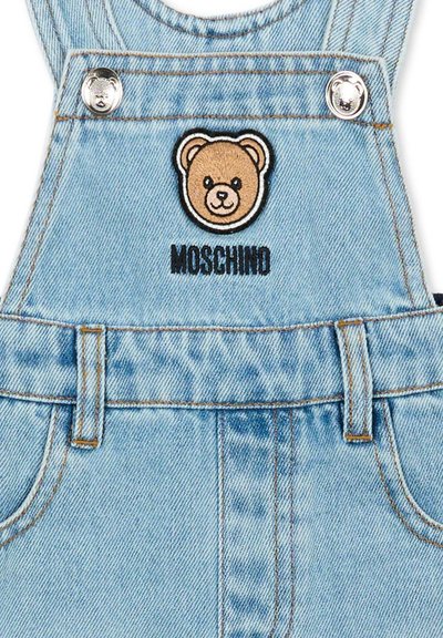 Jasnoniebieskie dżinsowe ogrodniczki z naszywką w kształcie twarzy misia i haftowanym napisem "MOSCHINO" poniżej, na obszarze kieszeni na piersi.