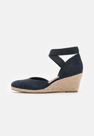 Marineblå ruskind kilehælssko med en espadrille-inspireret flettet jutesål og krydsede elastikstropper. Rundt tåparti, glat tekstur og afslappet design.