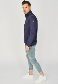 Laikku-zipup huppari tummansinisellä värillä, jossa on korkea kaulus, sivutaskut ja logo-ompelu hihassa, yhdistettynä kuluneisiin vaaleansinisiin farkkuihin ja valkoisiin lenkkareihin.