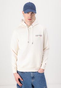 Hvit hettejakke med snor og frontlomme, med "TOMMY HILFIGER"-logo i rødt. Kombinert med blå caps og denimshorts.