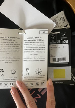 Etiquetas y rótulos de una prenda negra de O'Neill que indican talla 12, información sobre protección UV en italiano y francés, símbolos de cuidado y logotipos de certificación.