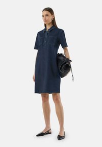 Denimkleid in Dunkelblau, knielang mit kurzen Ärmeln, mit geknöpftem Kragen und zwei Brusttaschen. Kombiniert mit einer schwarzen Handtasche und Ballerinas.