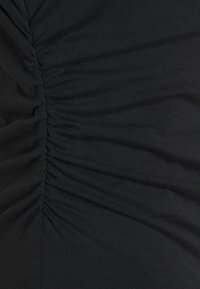 Haut en tissu extensible noir présentant une texture froncée et un discret détail froncé sur un côté. Finition lisse avec une qualité élastique.