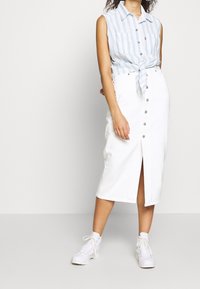 Kvinde iført en blå og hvid stribet ærmeløs knyttet skjorte, hvid knaplukket midiskørt og hvide snøresneakers, stående foran en ensfarvet baggrund.