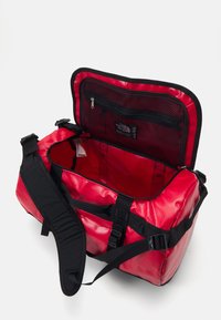 Red duffel bag with a black strap, zippered compartments, and a smooth, shiny texture. Interior includes a mesh pocket for organization.