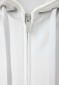 Gros plan d’une fermeture éclair d’un sweat à capuche blanc avec des cordons en tissu pendants de chaque côté de l’avant zippé.