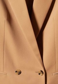 Blazer couleur camel avec larges revers, coupe croisée, deux boutons et poches fendues horizontales de chaque côté.