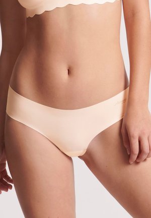 Frau trägt beige nahtlose Bikini-Unterwäsche und passenden BH, steht vor einem einfarbigen hellen Hintergrund.