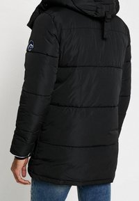 Giacca a vento nera con cuciture orizzontali, cappuccio e una patch con logo sulla parte superiore del braccio. Caratteristiche maniche lunghe e vestibilità comoda.