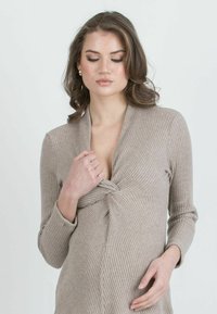Frau mit welligem braunem Haar, die ein beigefarbenes Rippstrickkleid trägt und einen vorderen Drehdetail hält, vor einem schlichten hellen Hintergrund.