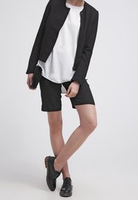 Svart kort blazer över en vit topp, kombinerad med svarta shorts. Modellen bär svarta snörskor och håller en liten svart clutch.