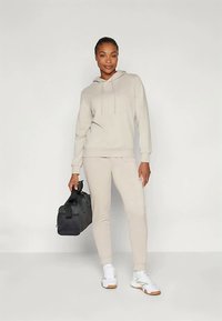 Žena v béžovej mikine a teplákových nohaviciach, s bielymi teniskami, drží čiernu športovú tašku a stojí pred jednoduchým svetlým pozadím.