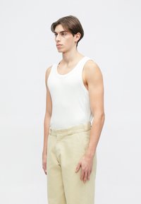 Biały prążkowany top na ramiączkach w połączeniu z jasnobeżowymi spodniami o luźnym kroju i prostym pasie, modelka stoi bokiem.