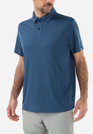 Homme portant un polo bleu à manches courtes avec col boutonné et pantalon gris clair, debout devant un fond blanc.
