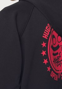 Sudadera negra con un gran diseño gráfico rojo que presenta una cara sonriente y estrellas. La tela parece suave con un estilo de capucha estándar.