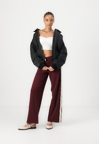 Veste bomber noire sur un top court blanc, accompagnée d'un pantalon large bordeaux avec une bande beige, et des chaussures de couleur claire.