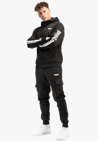 Melna zip-džemperis ar baltām akcentiem un logotipu uz piedurknēm, kombinēts ar saskaņotiem sporta biksēm, kas aprīkotas ar kabatām. Apavi ir baltas kedas.