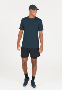 Leende man som bär svart keps, marinblå t-shirt, svarta shorts, svarta strumpor och marinblå löparskor, stående mot en enkel vit bakgrund.