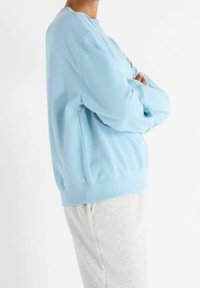 Person wearing a light blue sweatshirt and light gray pants, standing with arms crossed against a white background.