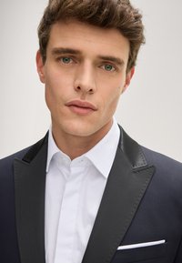 Giacca da smoking blu navy con revers in satin nero, fazzoletto da taschino abbinato e camicia bianca. Tessuto liscio, vestibilità su misura e dettagli nitidi.