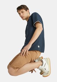 Camicia corta navy con bordi bianchi, tasca frontale; pantaloni corti beige; sneakers bianche con accenti grigi e suola in gomma; modello inginocchiato.