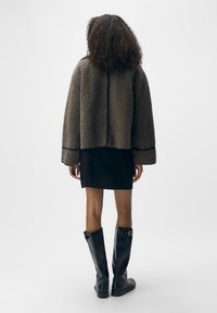 Giacca esterna marrone, morbida e pelosa, con una forma a scatola, indossata sopra un vestito nero corto; abbinata a stivali neri in pelle alti fino al ginocchio.