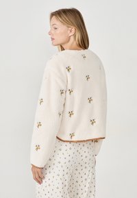 Kobieta z blond włosami ubrana w kremową polarową kurtkę z małym haftem kwiatowym i białe kwieciste spodnie, zwrócona w lewo na jednolitym tle.